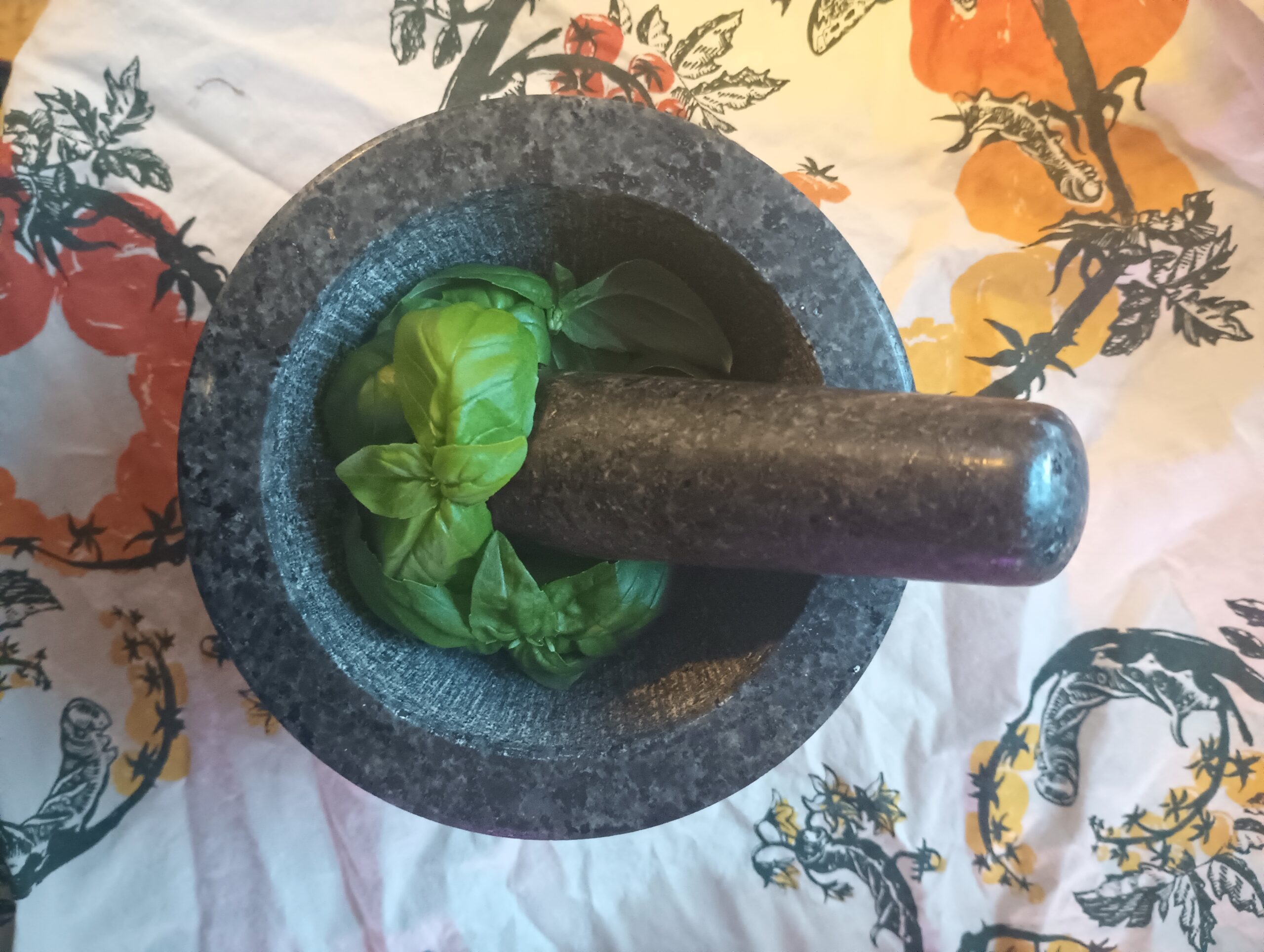 Siracusa basil tops next to a pestle, set in a mortar that is sitting on a white cloth screenprinted with illustrations of hornworms crawling on tomato plants