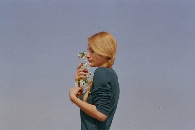 blond woman in green sweater smelling white flowers, sky background