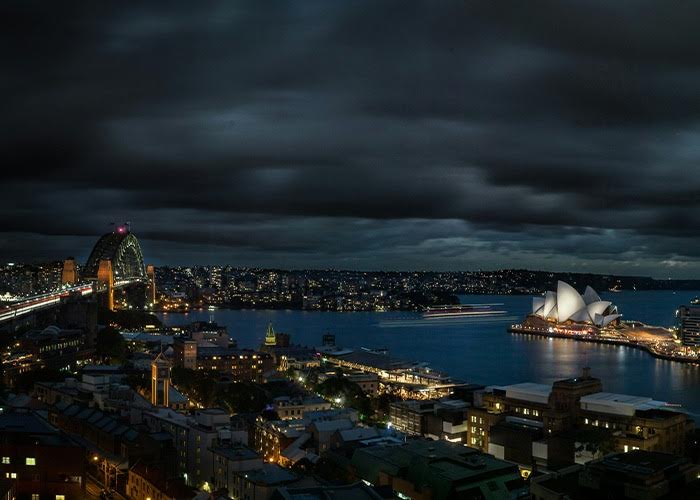 An aerial nighttime view of Sydney Harbour, with the Opera House’s glowing sails and the Harbour Bridge’s arch visible against the skyline